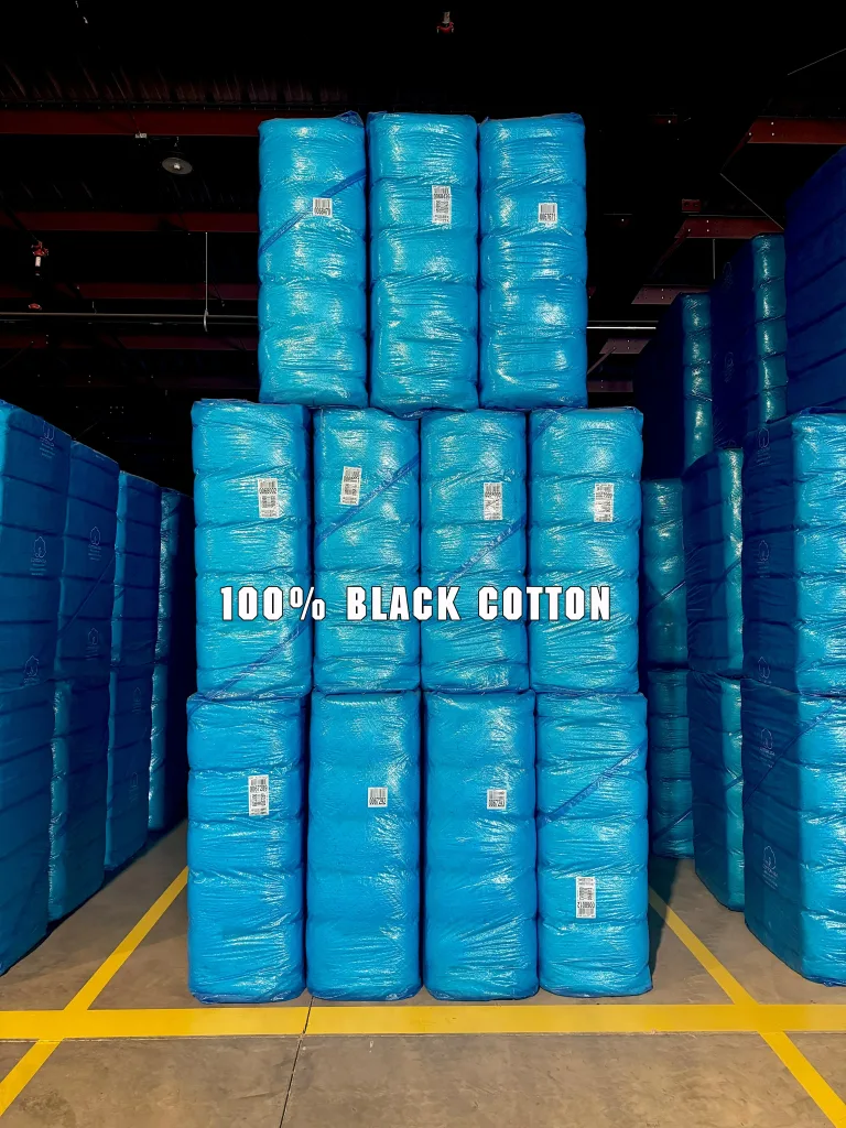 Stacks of blue-wrapped items arranged in a warehouse setting, with the text '100% Black Cotton' displayed prominently on the screen.