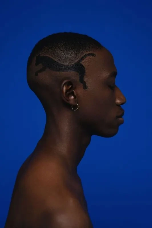 A close-up profile shot of a person with a distinct lion design shaved into their short hair against a vibrant blue background.