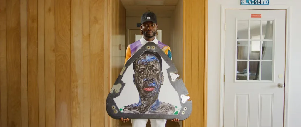 A man stands beside a door, holding a large shield with his painting of a black man's face on it, showcasing this artistic work and the entrance behind him.