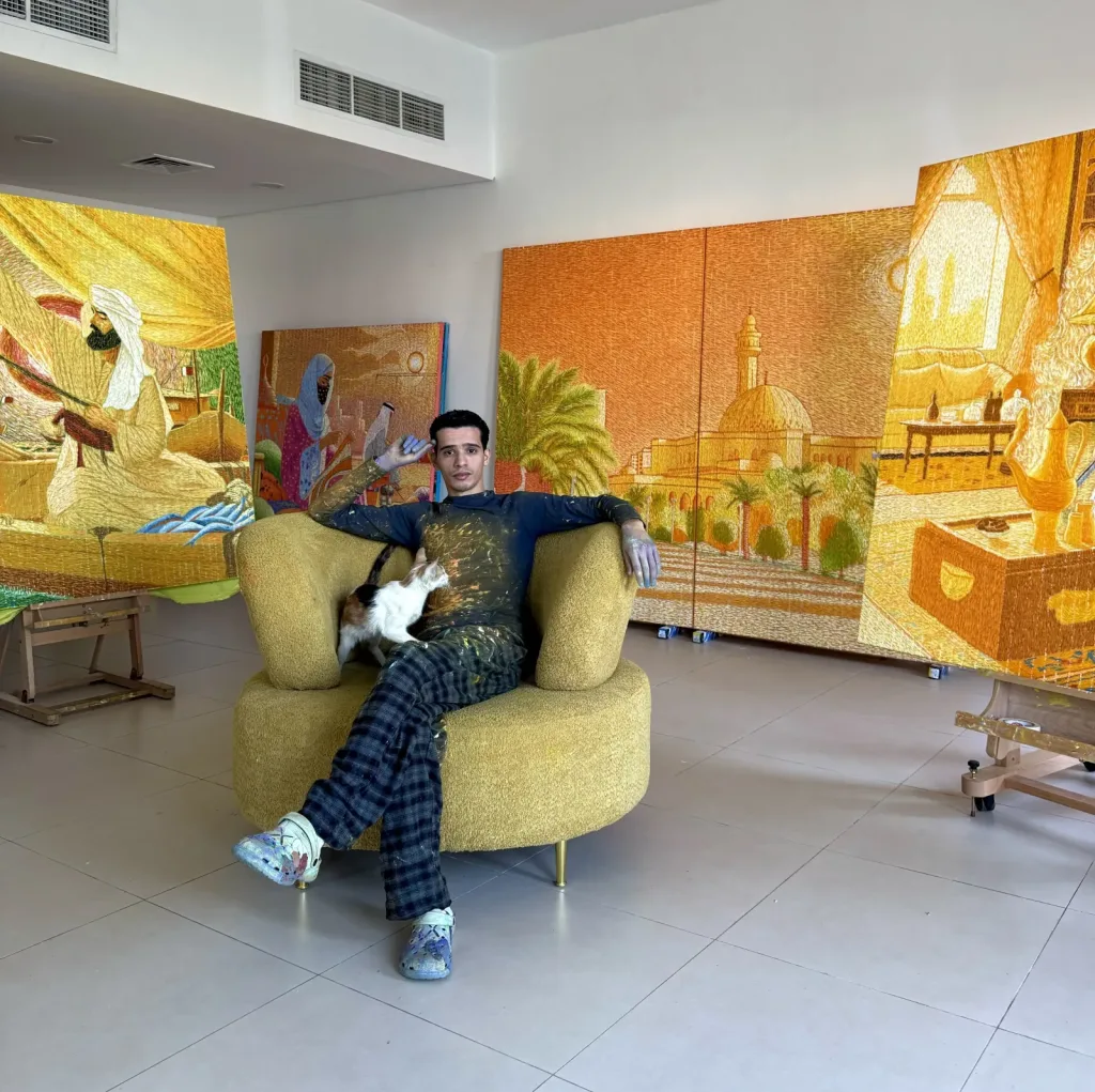 A person sits on a yellow chair with a cat on their lap, surrounded by large, colorful paintings depicting Middle Eastern scenes in a bright, modern room.