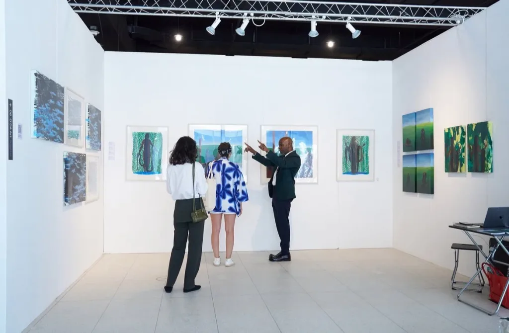 Three people in an art gallery viewing colorful paintings on white walls. The artwork features abstract and nature themes. The atmosphere is engaged and thoughtful.