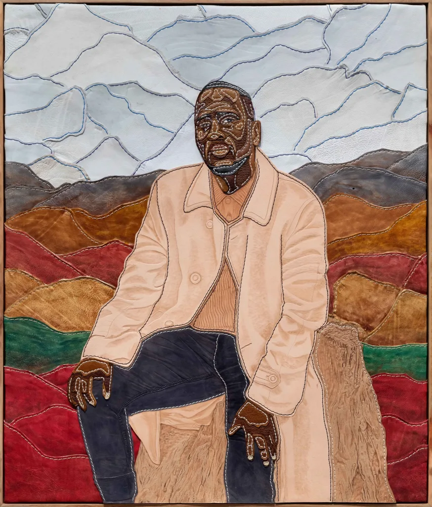Textile art depicting a person in a beige coat, seated against a backdrop of colorful stitched hills and a geometric-like sky.