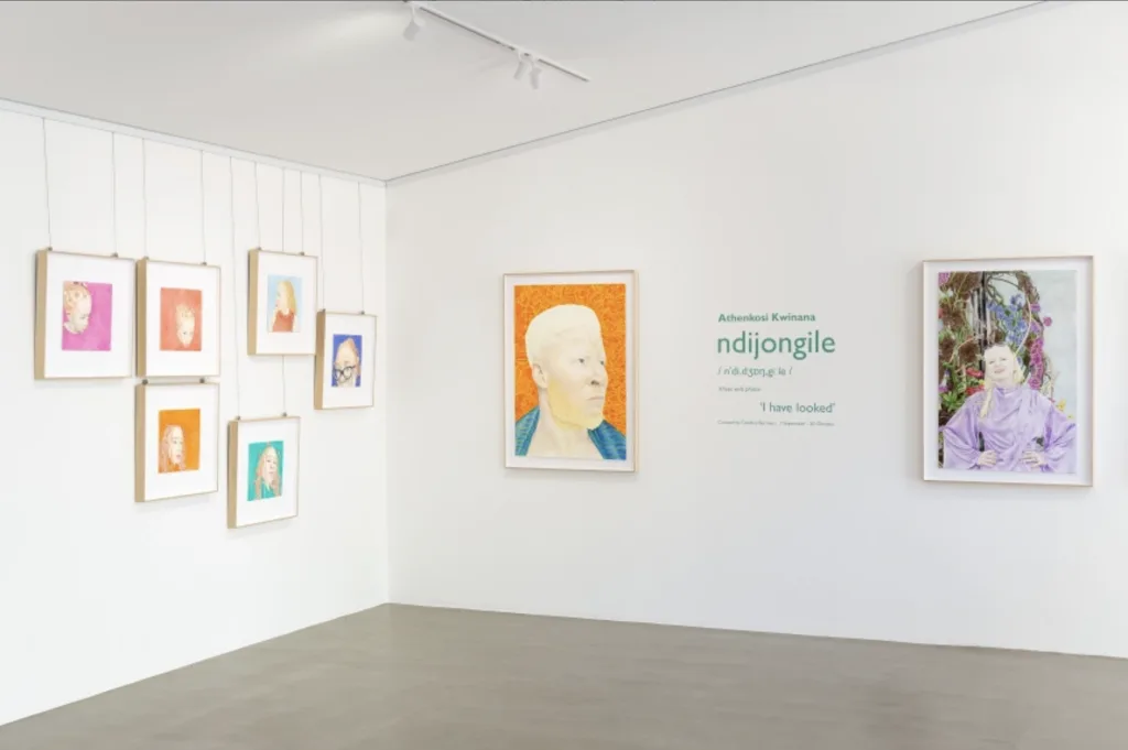 Art gallery showcasing portraits; left wall displays six varied framed pieces, right wall features two large colorful portraits with an exhibition title.