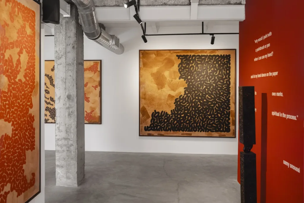 Art gallery interior with abstract paintings. Left: red patterns on tan; center: black shapes on gold. Red wall with white text. Concrete floor and gray ducts.