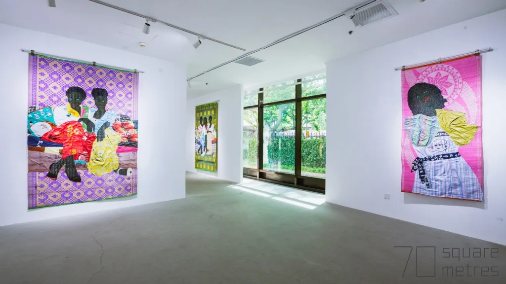 Bright art gallery with three colorful artworks on white walls. Large windows provide natural light and green outdoor views.