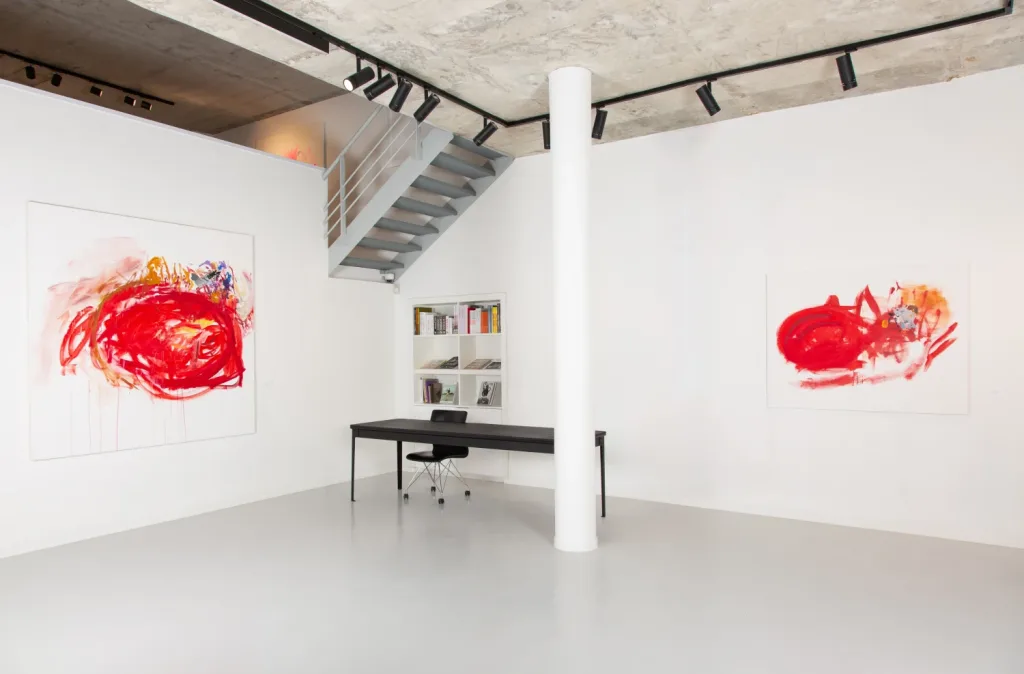 Minimalist gallery with two abstract paintings featuring bold red swirls on white walls. A sleek black desk and chair sit beneath metal stairs.