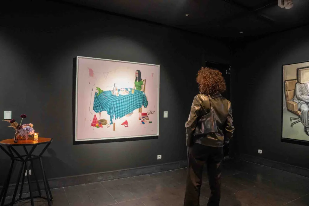 A person in a leather jacket observes a colorful painting of a woman at a table in a dimly lit art gallery. A small side table with flowers is nearby.